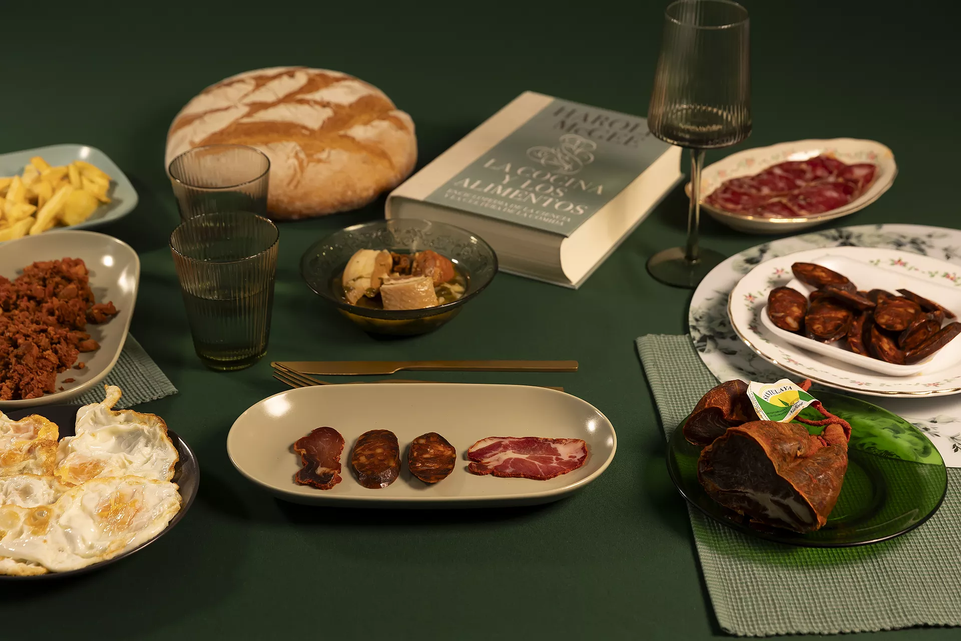 Bodegón de embutidos: chorizo, picadillo, lomo, chosco... en una mesa verde, junto a huevos fritos y una hogaza de pan